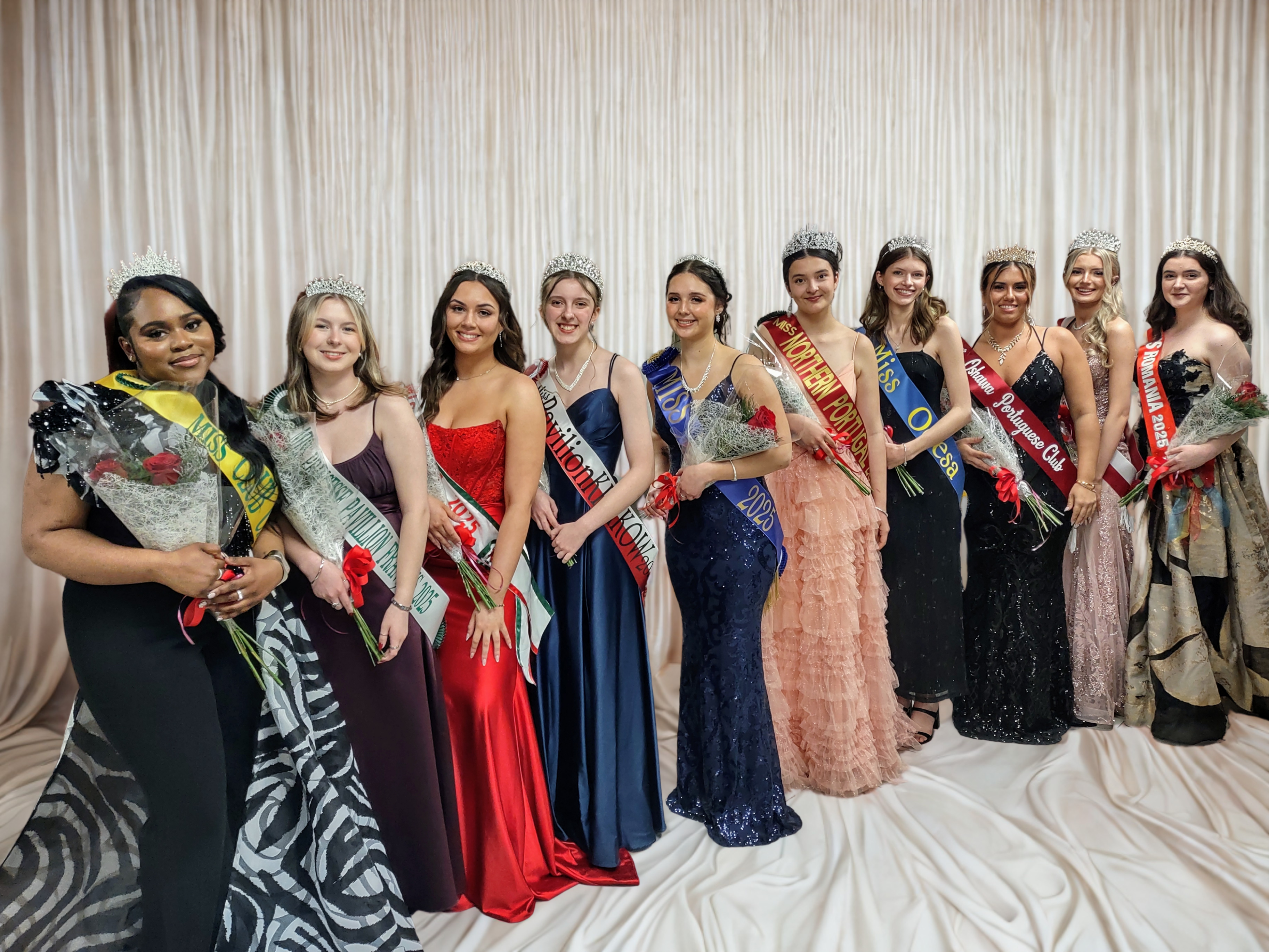 Ten young women stand a row. They are dressed in formal dresses and pant suits, with a sash, and a tiara on their heads. Each holds a bouquet of roses.